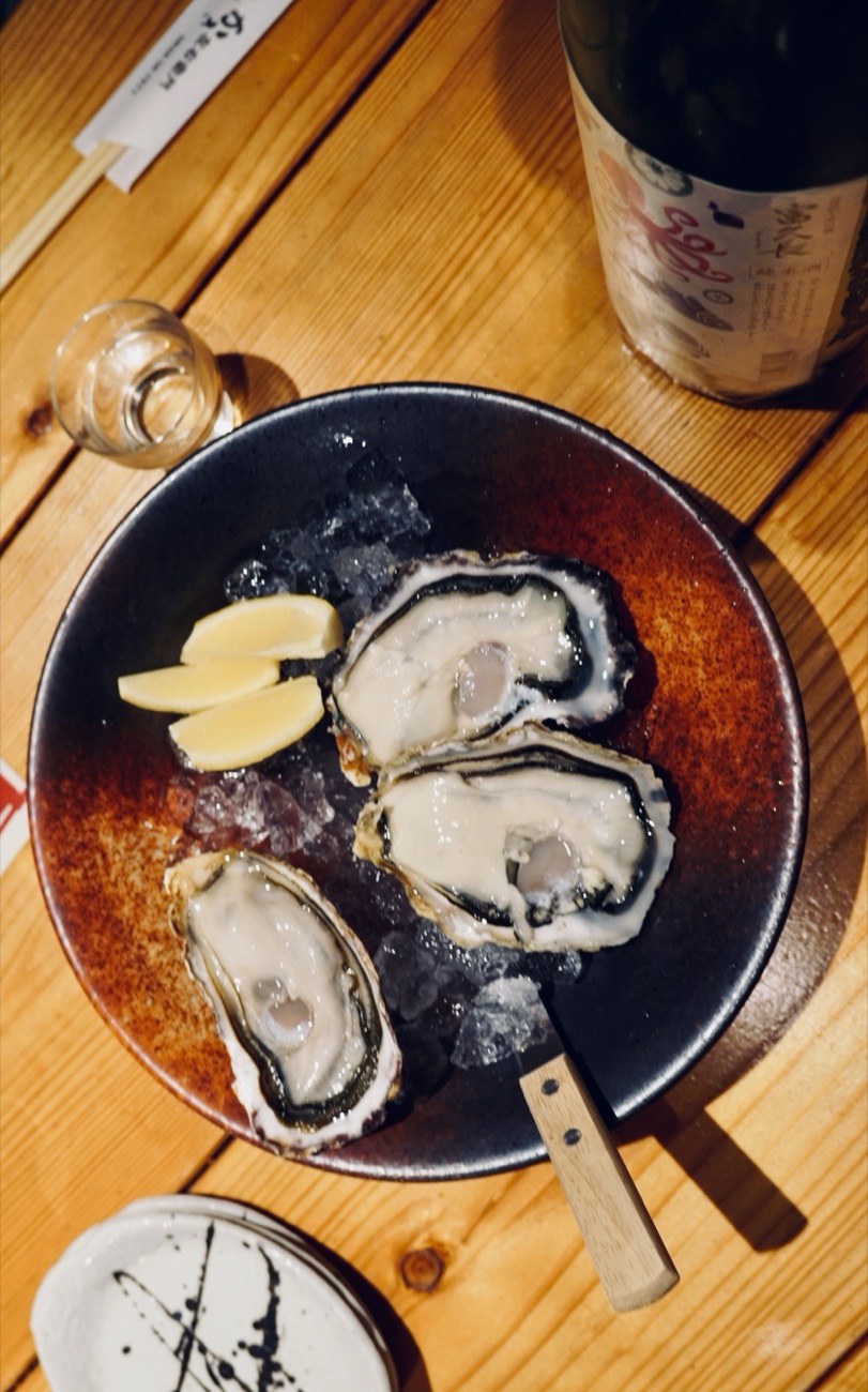 A bowl of fresh oysters on ice with lemon, beside a bottle of sake